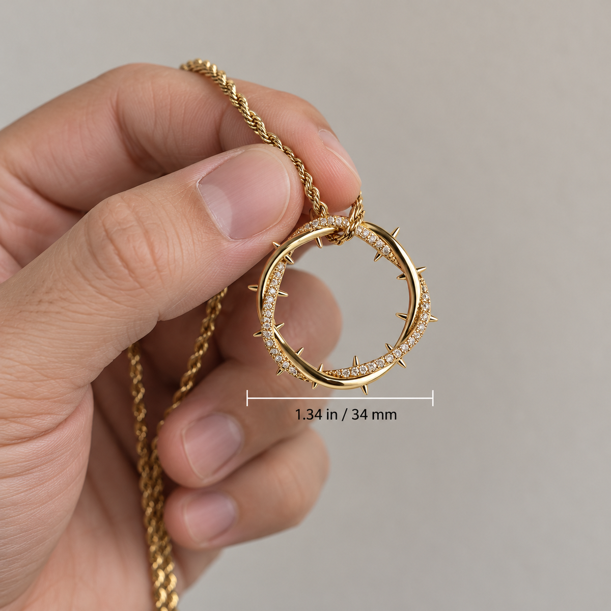 Hand holding the gold crown of thorns pendant between two fingers to show its 1.34-inch size, with the chain draped over the hand.