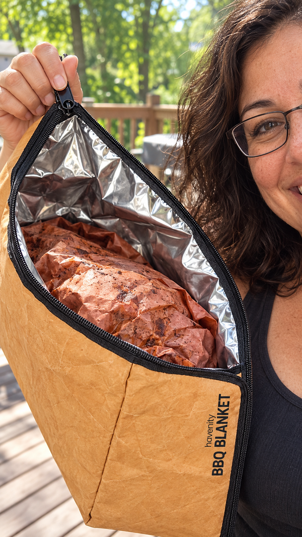 A woman holds the BBQ Blanket open toward the camera, showing the foil lining and a wrapped pork shoulder inside.