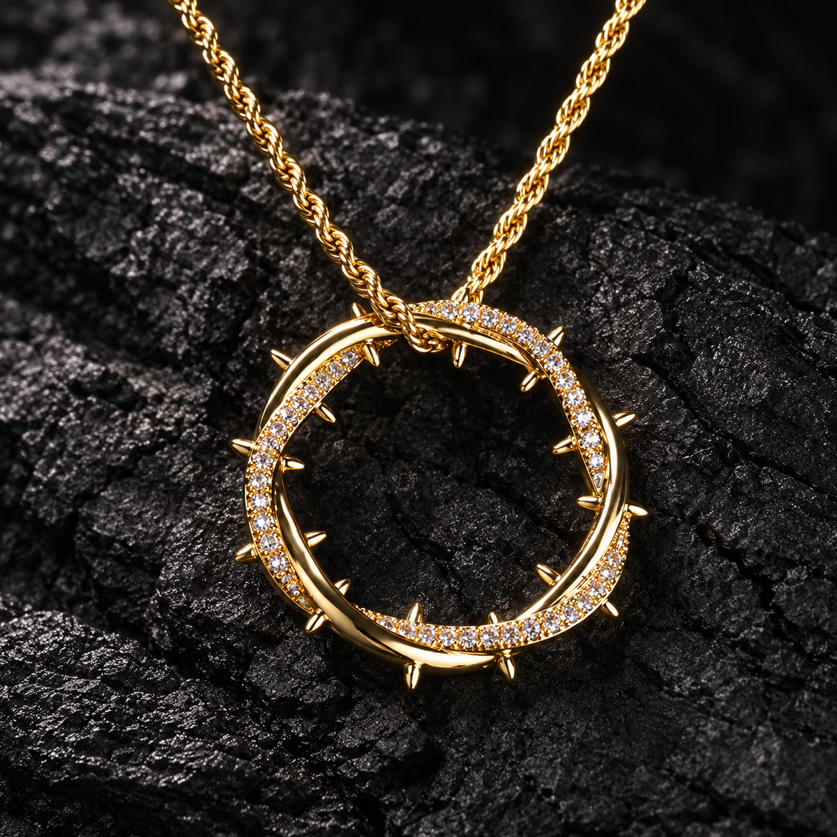 Gold crown of thorns pendant with clear CZ stones on a gold rope chain, lying on dark textured stone.