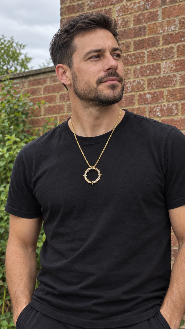 A bearded man in a black T-shirt wears the gold Crown of Thorns Necklace in front of a brick wall.