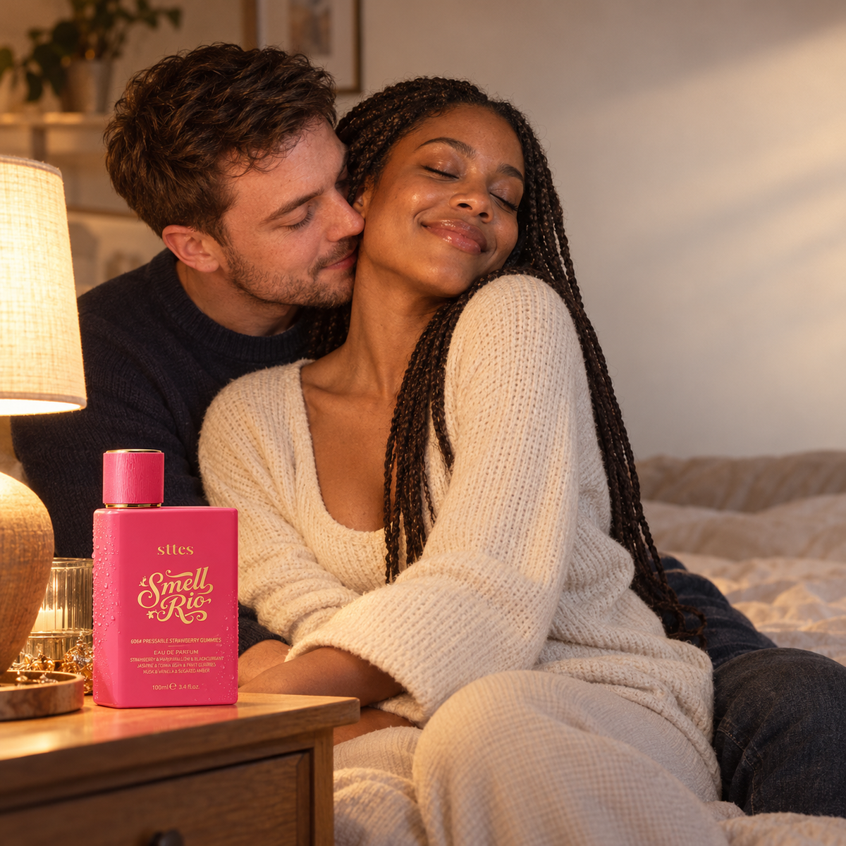 A young man leans in to smell a woman's hair in a warmly lit bedroom, with the pink Strawberry Gummies perfume bottle on the nightstand.