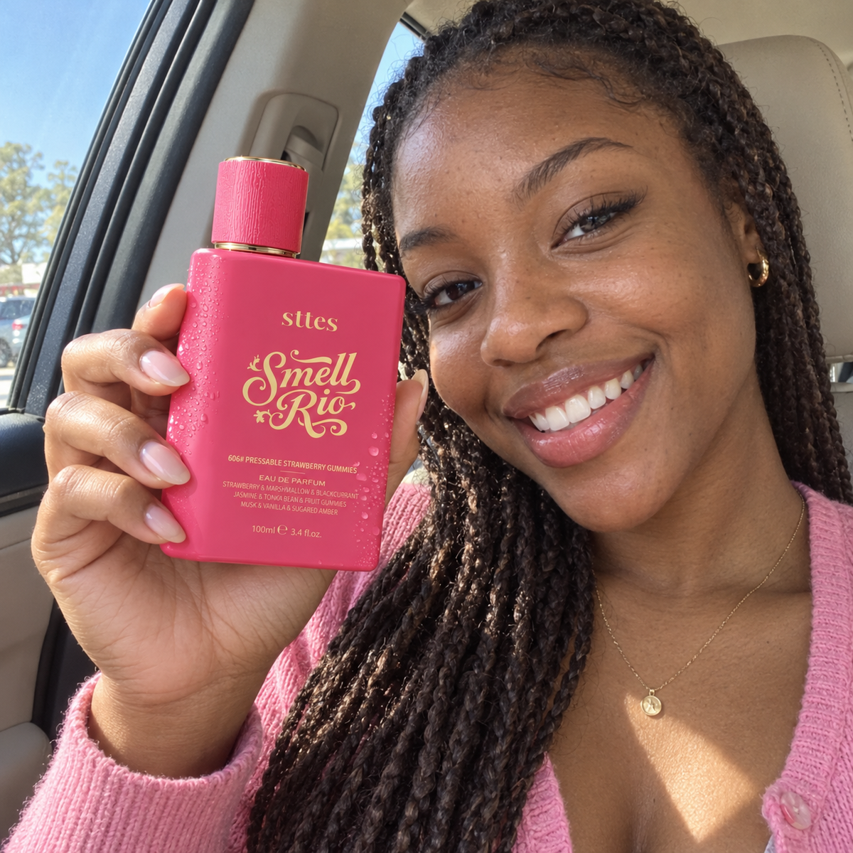 A smiling young woman in a car holds the pink Strawberry Gummies perfume bottle up beside her face.